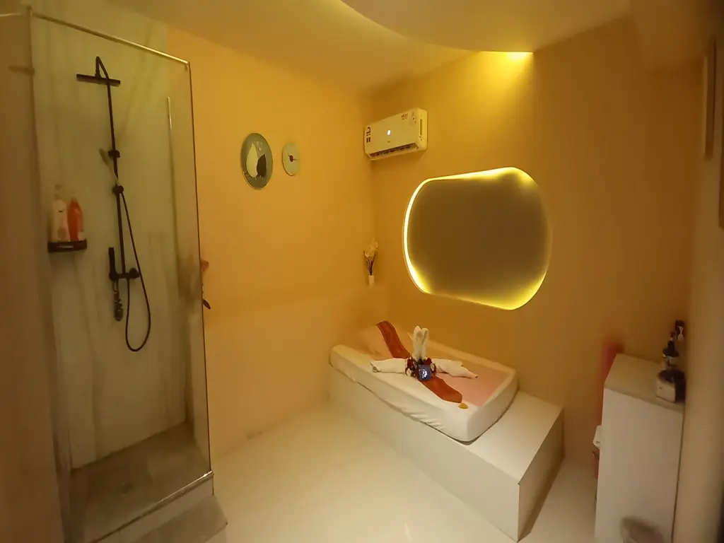 Hotel bathroom with a raised bed platform, warm yellow lighting, and a round backlit mirror; glass shower on the left and a small cabinet on the right.